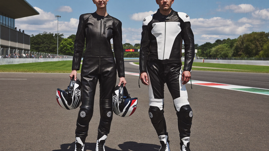Two motorcycle riders comparing one-piece and two-piece riding suits on a sunlit racetrack